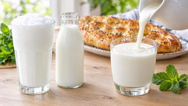 A frothy glass of Turkish ayran and a bottle of ayran next to a glass of thick kefir, with bread and mint